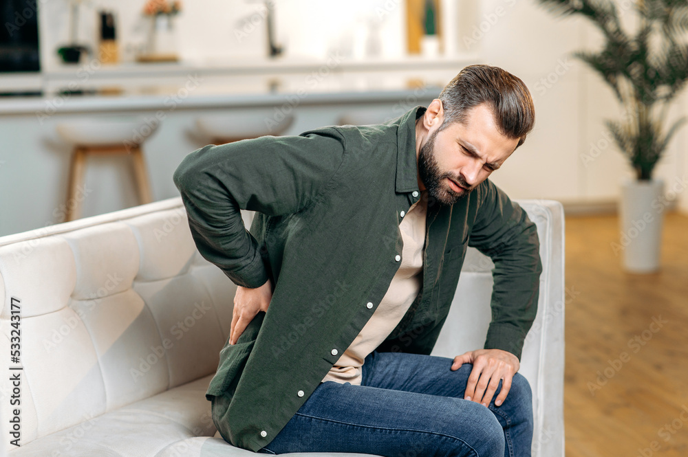 Unhappy caucasian man, dressed in casual wear, sitting on sofa in living room, experiencing acute pain in the back, lower back, has chronic joint problems. Problems with the back, spine, sciatica