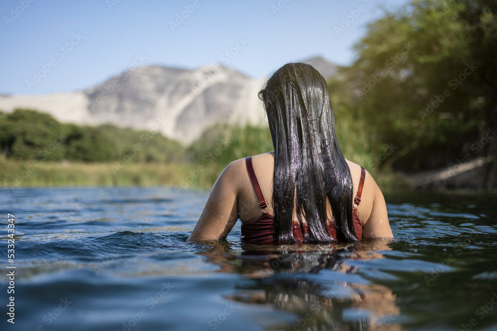Fototapeta premium Beautiful young latin woman bathes in a lake enjoying a summer day. It stands at the bottom of the shoulders in the water.
