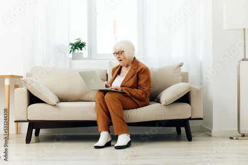 a sweet elderly woman with gray hair is sitting on a beige sofa in a brown suit working remotely at a laptop and lifting her glasses from her face, looking intently at the monitor