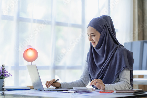 Asian Muslim businesswoman in hijab using laptop and working with charts, financial documents, analyzing accounting calculations at office.