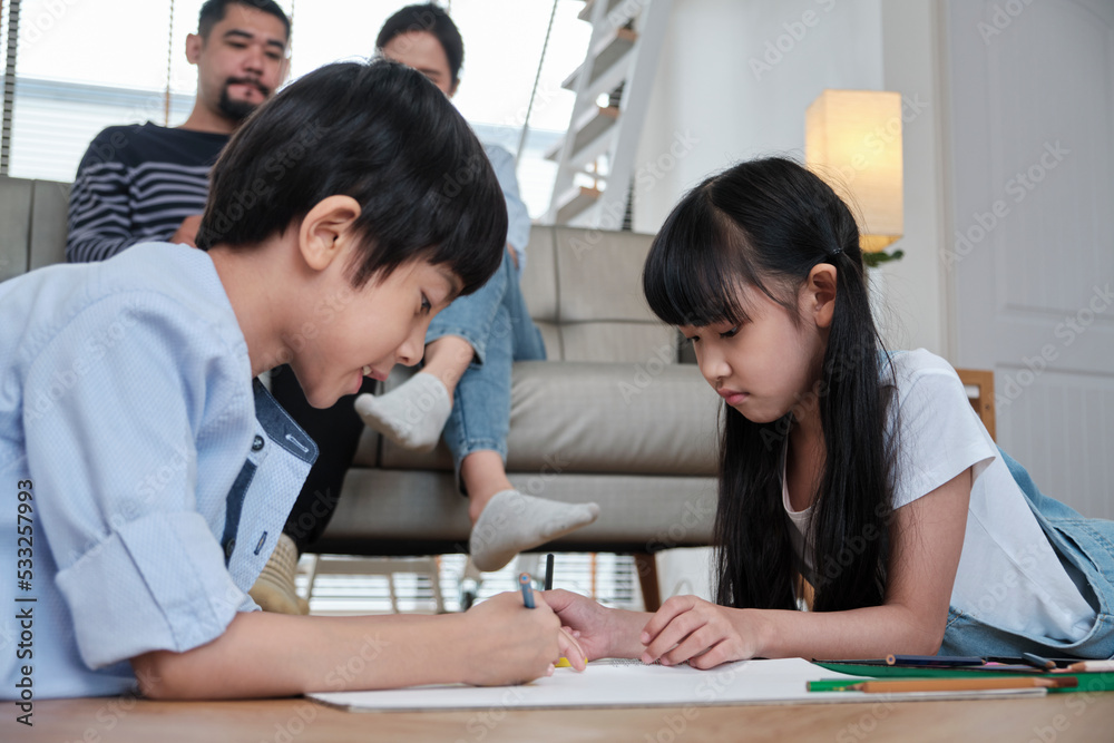 Asian Thai siblings are lying on living room floor, drawing homework with colored pencils together, parents leisurely relax on a sofa, lovely happy weekend activity, and domestic wellbeing lifestyle. Asian Thai siblings are lying on living room floor, drawing homework with colored pencils together, parents leisurely relax on a sofa, lovely happy weekend activity, and domestic wellbeing lifestyle.