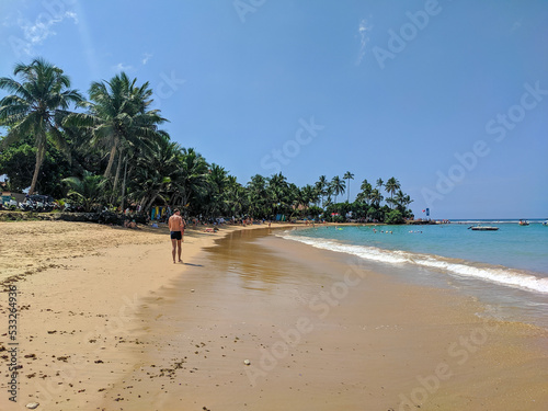 Hikkaduwa, Sri Lanka - March 8, 2022: Beautiful view of the beach in Hikkaduwa with green palm trees. People relax, sunbathe and swim in the Indian Ocean