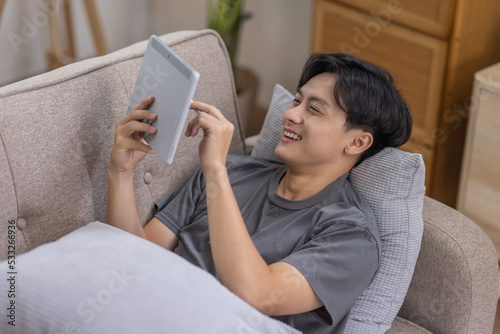 Handsome Asian man using digital tablet while headphones sitting on sofa at home