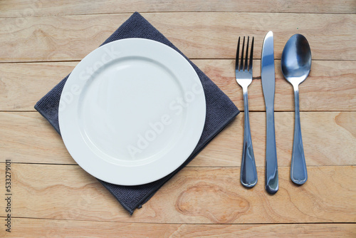 Empty white plate on cloth with fork, spoon and, knife, tableware isolated on wooden background closeup.
