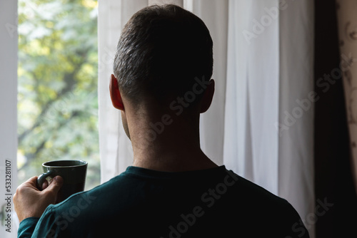 Back of young adult man with cup of coffee near window