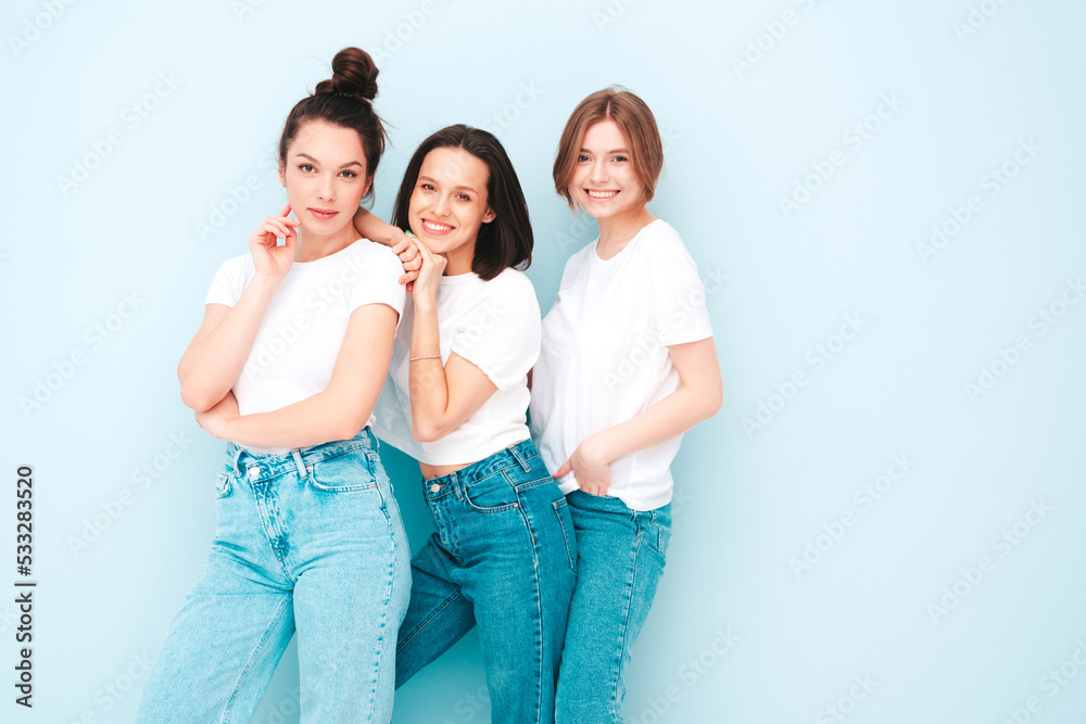 Fototapeta premium Three young beautiful smiling hipster female in trendy same summer white t-shirt and jeans clothes. Sexy carefree women posing near light blue wall in studio. Cheerful and positive models having fun