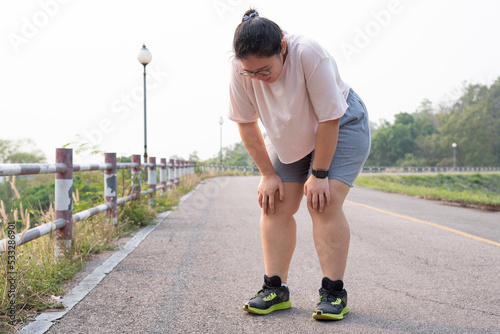Wide shot portrait of overweight Asian woman wearing eyeglasses, standing outdoor, bending down, resting after running, being tired, suffering from knee and joint injury. Sport and obesity concept.