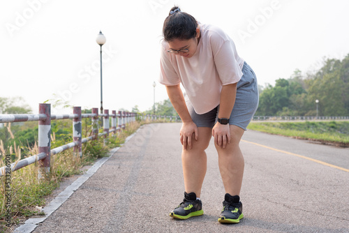Quadro em tela Wide shot portrait of overweight Asian woman wearing eyeglasses, standing outdoor, bending down, resting after running, being tired, suffering from knee and joint injury