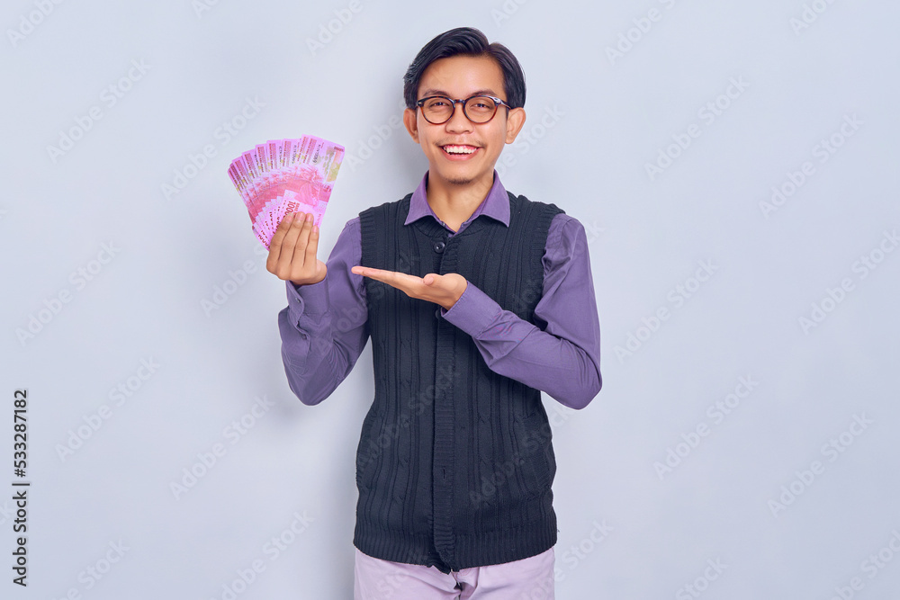 Portrait of smiling young handsome Asian man in shirt clothes showing cash money in rupiah banknotes with hands, looking at camera isolated on white background. People lifestyle concept Portrait of smiling young handsome Asian man in shirt clothes showing cash money in rupiah banknotes with hands, looking at camera isolated on white background. People lifestyle concept