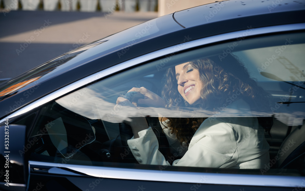Beautiful businesswoman with closed eyes sitting in electric car and smiling while using vehicle autopilot system. Pretty woman holding steering wheel and sleeping while sitting on driver seat in car.