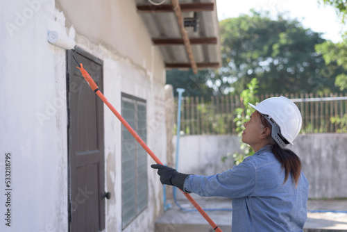Side view medium shot of a senior Asian woman wearing safety hard hat, gloves, holding paint roller with extension handle, painting old wall, concentration. Retired, worker, DIY, renovation concept.