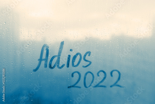 lettering Adios in Spanish is goodbye in english and numbers 2022 paint with finger of water on splashed by rain foggy glass on sunset window