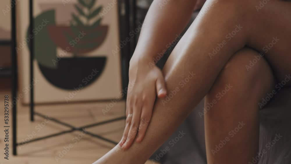 Attractive young African American woman with tattoo on chest sitting on bed in morning applying and dabbing body cream over her legs
