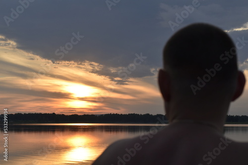 A man stands with his back against the background of a beautiful sunset sky over the lake.