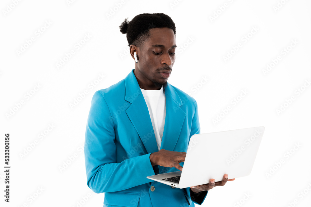 Serious african american man sitting on floor on white background in studio isolated looking at screen holding modern laptop listening to music by modern earphones. Serious african american man sitting on floor on white background in studio isolated looking at screen holding modern laptop listening to music by modern earphones.