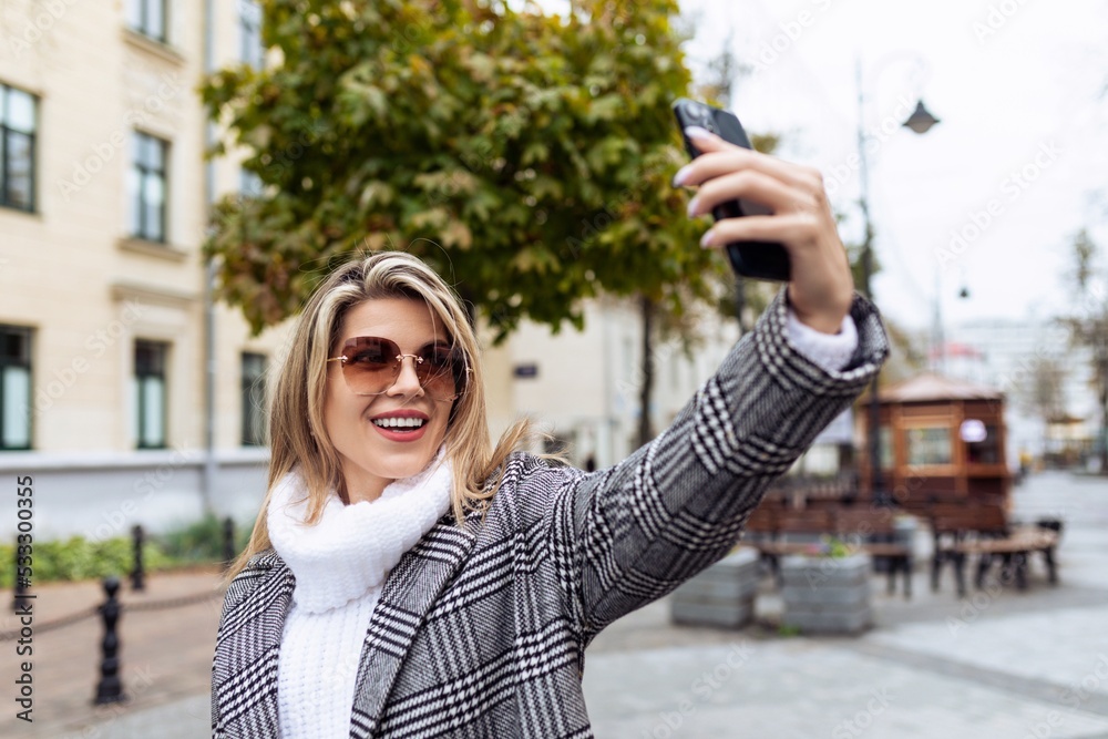 an adult woman in a white sweater and sunglasses makes a selfie on the phone on the background of the city