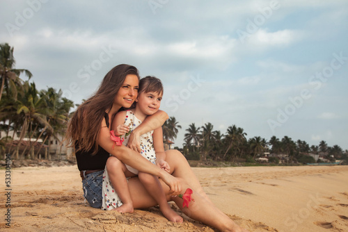 Mother and little daughter together sitting on tropical sandy beach at palm trees and sky background. Mom and little girl in casual clothes on sea. Concept family vacation, travel. Copy text space