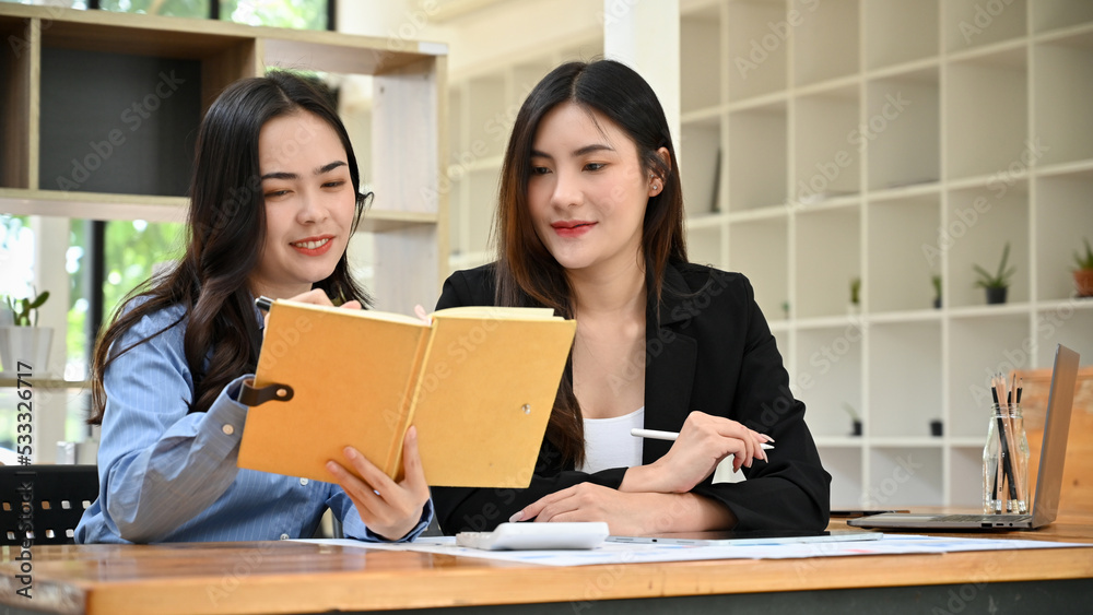 Asian businesswomen working together in the office, looking for information in a book. Asian businesswomen working together in the office, looking for information in a book.