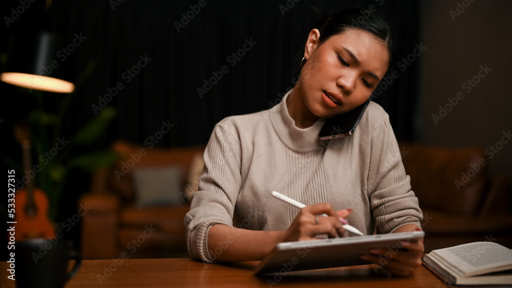 Asian businesswoman lists some details on a tablet while on the phone with a client Asian businesswoman lists some details on a tablet while on the phone with a client
