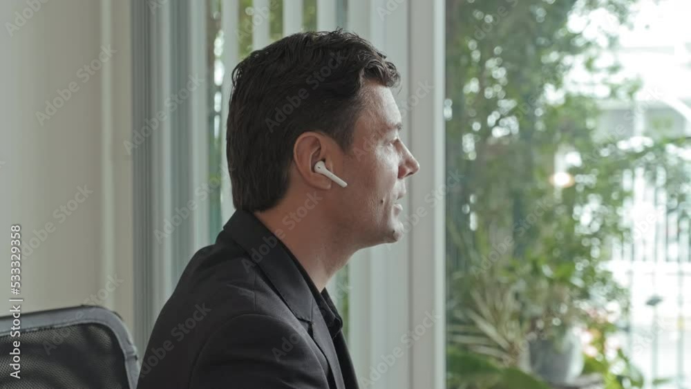 Chest up shot of successful businessman in wireless earphones having video call with his team on computer, sitting at desk in his office with panoramic windows Chest up shot of successful businessman in wireless earphones having video call with his team on computer, sitting at desk in his office with panoramic windows