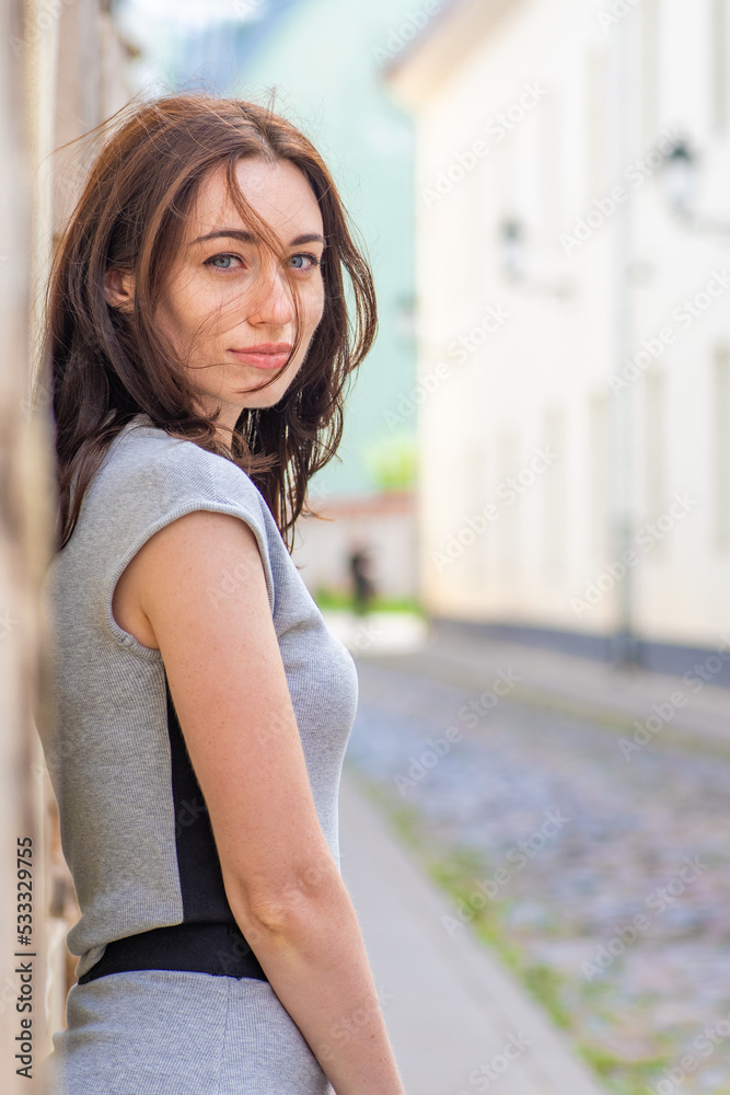 Beautiful Ukrainian girl with red hair, blue eyes and freckles in a grey summer dress leaning with his back against a wall in a cobbled street with street lamps in the old town, vertical