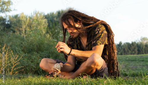 Young man with long dreadlock hitting stone on stone to make a primal fire.  Survival in nature concept.