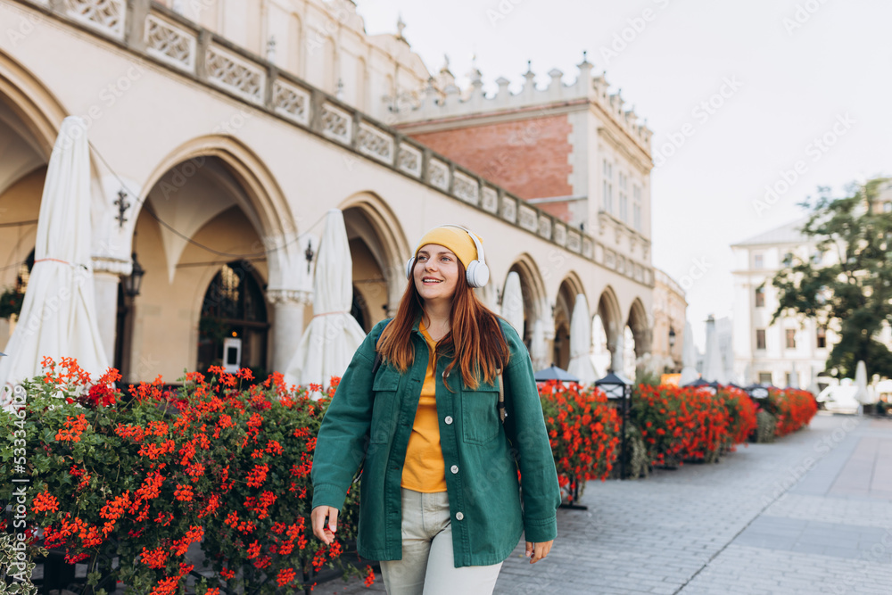 Young happy woman traveler in yellow hat listening to music through wireless earphones on urban background. Stylish female foreigner examines architectural monument during her long-awaited vacation