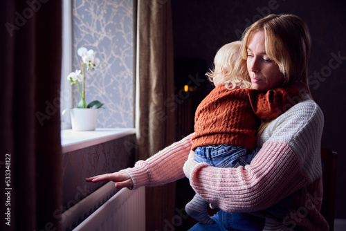 Mother With Son Trying To Keep Warm By Radiator At Home During Cost Of Living Energy Crisis photo
