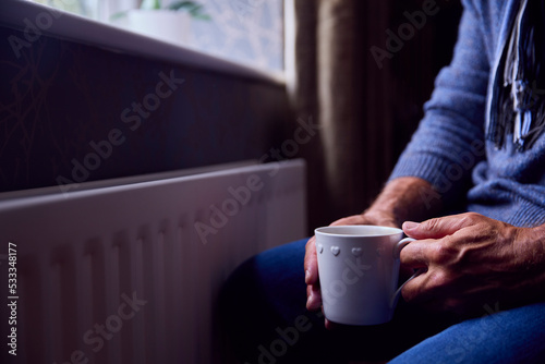 Close Up Of Mature Man Trying To Keep Warm By Radiator At Home During Cost Of Living Energy Crisis