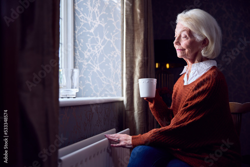 Senior Woman With Hot Drink Trying To Keep Warm By Radiator At Home In Cost Of Living Energy Crisis