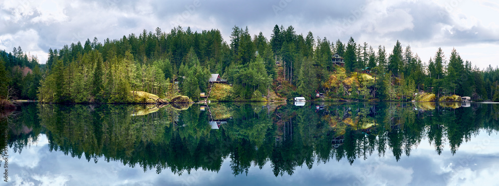 Fototapeta premium Houses on the rocks among the coniferous forest are reflected in the crystalline water of the wild Ruby Lake in calm weather. Forest landscape at Dan Bosch Park, Sunshine Coast Hwy, BC, Canada