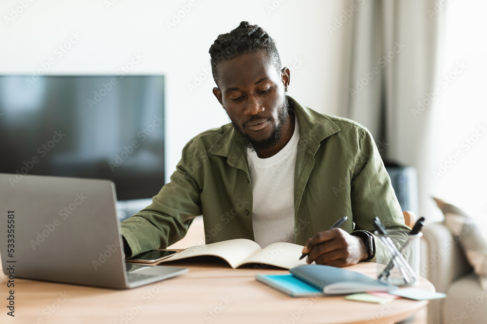 Focused african american man sitting at desk working on laptop and taking notes in notebook, studying online at home Focused african american man sitting at desk working on laptop and taking notes in notebook, studying online at home