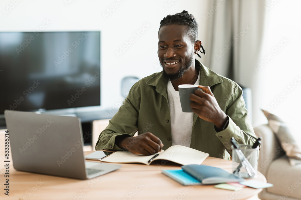 Happy black man drinking cup of tea at workplace while working on computer in home office interior Happy black man drinking cup of tea at workplace while working on computer in home office interior