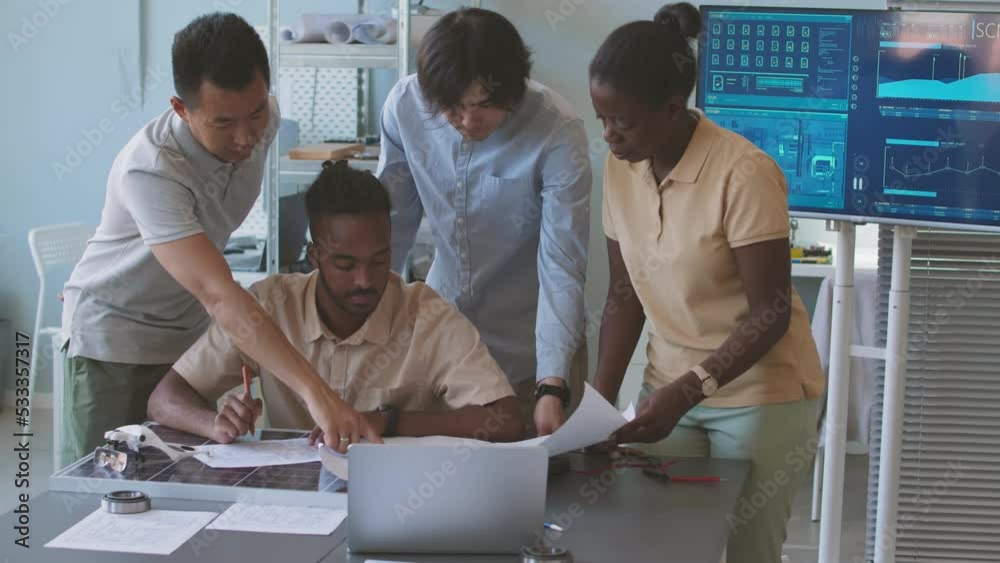 Four diverse electrical engineers discussing technology schemes while developing environmental project together in office, working at table with solar panel on it