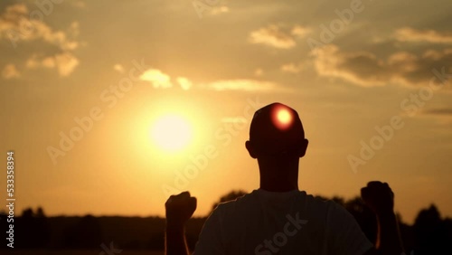 The silhouette of a man at sunset stands with his hands raised with happiness photo