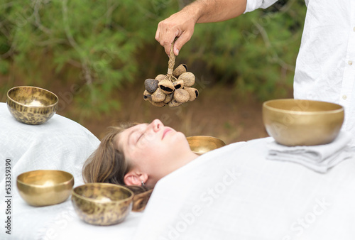 Male hand holding a Seed shell shaker musical instrument on sound healing therapy with tibetan singing bowls over young woman during healing sound massage outdoor, selective focus