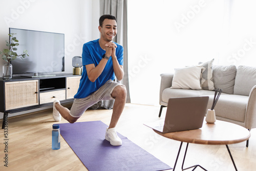 Happy arab man doing forward lunge exercises while watching online workout tutorial via laptop, training at home