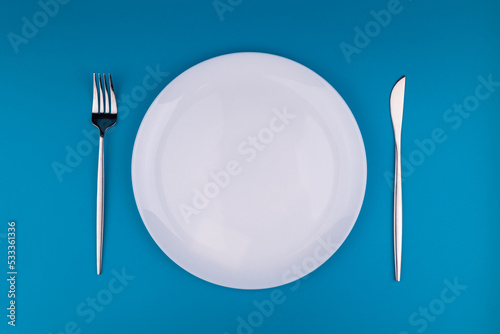 Stylish table setting, empty white plate with fork and knife on blue background, top view, flat lay