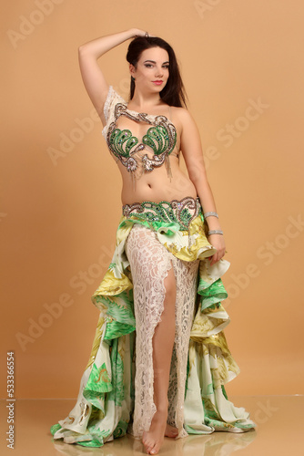 a pretty girl brunette who dances go-go and belly dance, stretching posing in studio in green arabian dress on a beige background