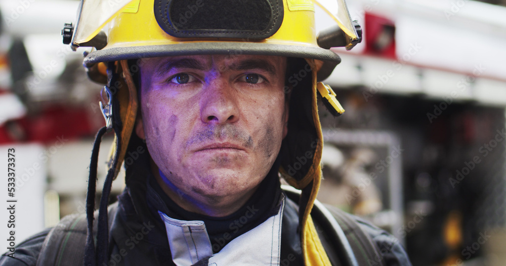 Close up portrait of handsome fireman in helmet and gull equipment standing next to the car with flashing lights on and looking into camera. Concept of saving lives, heroic profession, fire safety Close up portrait of handsome fireman in helmet and gull equipment standing next to the car with flashing lights on and looking into camera. Concept of saving lives, heroic profession, fire safety