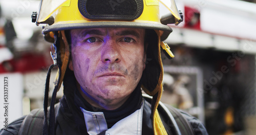 Close up portrait of handsome fireman in helmet and gull equipment standing next to the car with flashing lights on and looking into camera. Concept of saving lives, heroic profession, fire safety