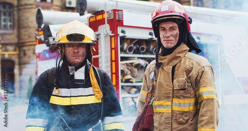 Photos Medium plan of two firefighters in helmet and gull equipment standing next to car and looking at camera