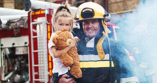 Medium plan of firefighter approaches from fire van with flashing lights on with cute girl with teddy bear. Smoke from fire covers rescuers. Concept of saving lives, heroic profession, fire safety photo