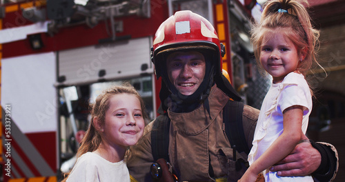 Portrait of the handsome young Caucasian fire fighter holding two small saved girls in hands while they looking at camera and happy smiling being outdoors next to red big fire truck background.