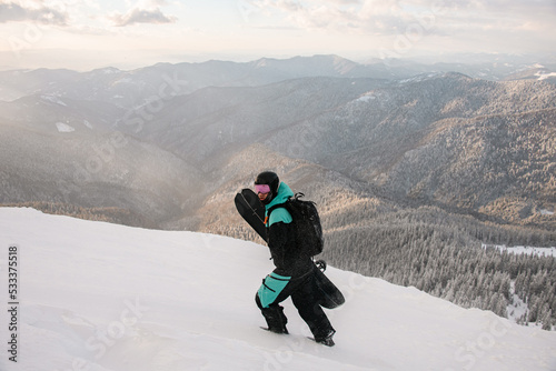 tourist with snowboard walks at snow-covered slope against the backdrop of mountain landscape