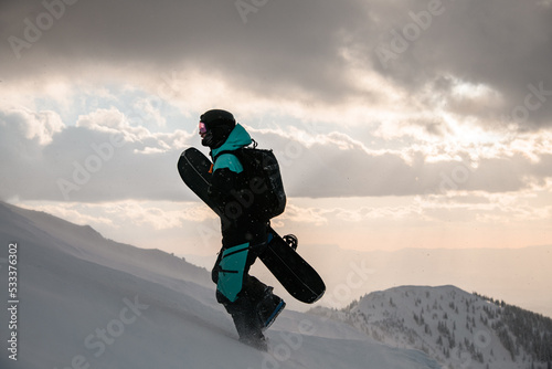 side view of man in ski suit with snowboard walks at snow-covered slope