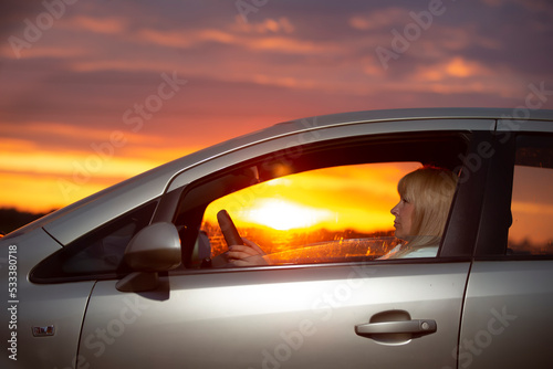 A middle-aged woman driving a car against the backdrop of a sunset sky.