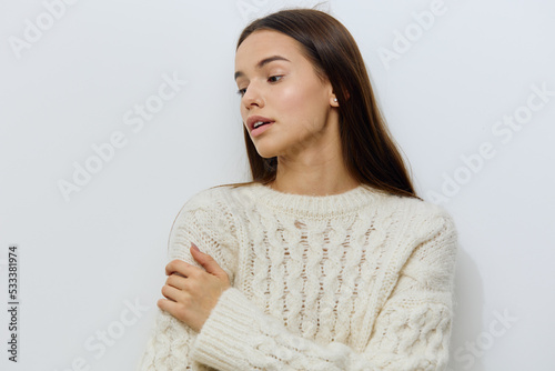 horizontal photo of a beautiful, gentle young woman, brunette standing in a white, warm, stylish sweater on a light background, with her hand on her shoulder and looking away