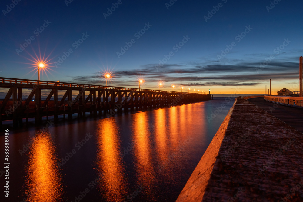 Hafenausfahrt Blankenberge zum Meer mit einer langen Holzbrücke und beleuchteten Laternen in der Abenddämmerung 