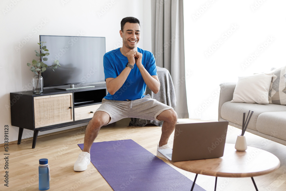 Young arab man doing deep squats, looking at laptop during online training, exercising at home, free space Young arab man doing deep squats, looking at laptop during online training, exercising at home, free space